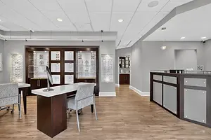 The interior of an optometrist’s office has a wooden floor and a white desk with chairs and a monitor.