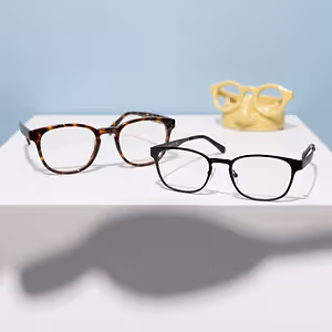 Three pairs of glasses sit on a white surface, including a brown pair, a black pair, and a yellow pair.