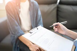 A person is holding a clipboard with a pen and paper while another person is sitting beside them and holding a clipboard with a pen.