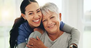 A woman with gray hair is smiling and hugging an older woman with a gray t-shirt. They are both standing in front of a white wall.