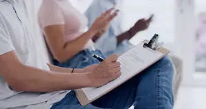 A man writes on a clipboard while sitting on a couch with two other people. The man is wearing a gray shirt and blue jeans. The two other people are sitting behind him, and they are both looking at their phones. The man is looking at the paper he is holding while holding a pen in his hand. Behind him is a glass window with a white frame.