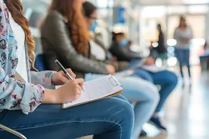 A woman is writing on a clipboard in a hospital waiting room with other people wearing face masks.