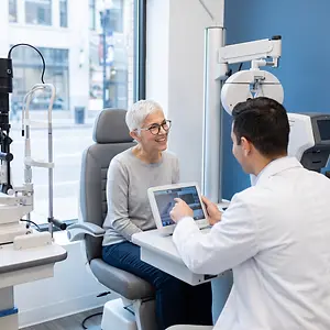 A woman wearing glasses and a gray sweater sits in an optometrist's chair while a man in a white coat examines her with a tablet