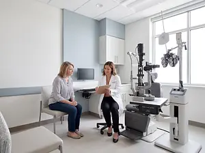 A woman in a white coat is sitting in a chair and holding a clipboard while another woman in a chair is looking at her.