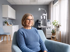 A smiling woman with short hair wearing glasses and a blue sweater sits in a chair in a clinic.