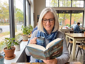 A woman with white hair and glasses sits at a window table with a cup and a book titled The Covenant of Water.