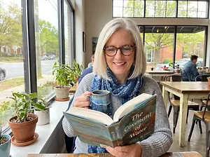 A woman with white hair and glasses sits at a window table with a cup and a book titled The Covenant of Water.