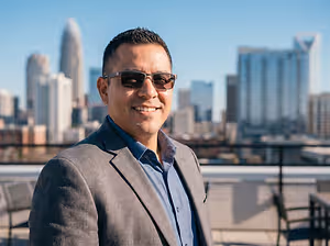 A man wearing sunglasses and a suit jacket standing on a rooftop in front of a cityscape.
