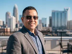 A man wearing sunglasses and a suit jacket standing on a rooftop in front of a cityscape.