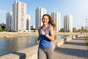 A woman is running and smiling in front of a river with buildings in the background.