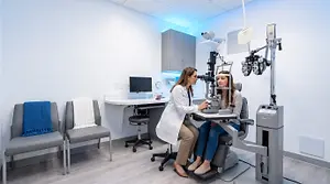 A woman is sitting in front of an eye exam machine while another woman stands in front of her with an eye exam machine and a monitor behind her.