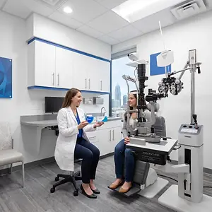 Two women sitting in a modern exam room, one of them is holding an eye exam card and the other one is sitting on the eye exam chair.