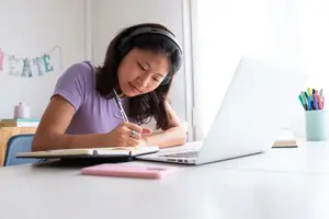 A woman sitting on a chair wearing headphones is writing on a notebook while looking at a laptop