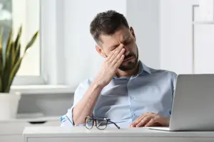 A man wearing glasses and a shirt is sitting in front of a laptop and touching his forehead.