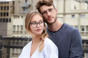 A man and a woman wearing glasses are standing on a balcony. They are both looking forward and posing for a photo. Behind them is a metal fence and a building with glass windows.
