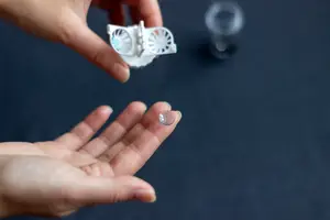 A hand holding a white contact lens case with a lens inside and a glass on a table
