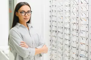 A woman in a white shirt and glasses stands in front of a wall of eyeglasses, smiling confidently.