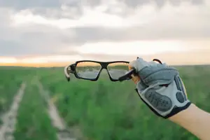 A hand holds glasses with a reflective surface in a grassy field.
