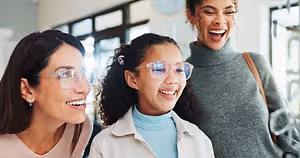 Three women are standing together and smiling at each other. The woman on the left is wearing glasses and earrings. The girl in the middle is wearing glasses and a turtleneck shirt. The woman on the right is wearing a turtleneck shirt, a gray sweater, and carrying a handbag.