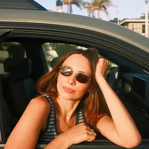 A woman with brown hair wearing sunglasses is leaning out of a car window with her hand on her head.
