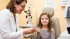 A young girl smiles as an eye doctor examines her eyes with a device in an eye care clinic.