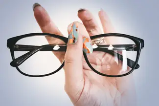 A hand holds a pair of black glasses with clear lenses, fingers showing colorful nail polish.