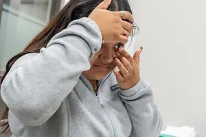 A woman wearing a gray jacket, multiple rings, and a necklace is holding her head and face with both hands, possibly in a medical setting.