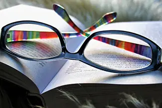 A pair of colorful eyeglasses with a black frame rests on top of an open book with visible text and a white page, set against a blurred background.