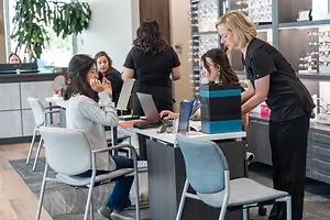 A group of women are in a room with a woman in a black shirt standing next to a desk with a laptop, a chair, and a box. The woman in the black shirt is probably helping the woman in the white shirt, who is sitting on a chair and is probably applying makeup. Behind them is a shelf with glasses on display. On the left side, there is a plant and a glass window with a view of the outside.