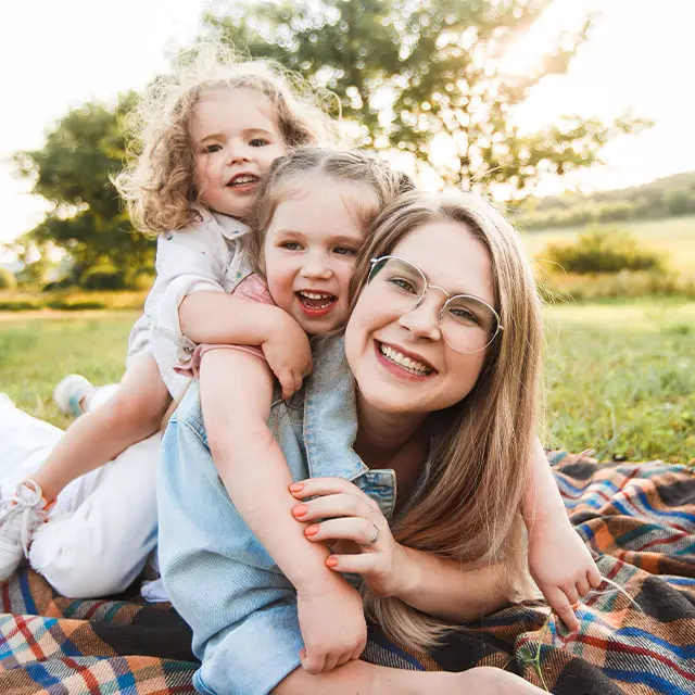 Two young girls and their mother sitting on a blanket on a grassy field, all smiling