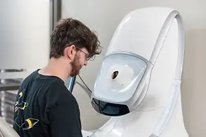 A man wearing glasses is sitting in front of a white machine, possibly a robotic arm, and is looking at a monitor that displays a graph. He is wearing a black shirt with a yellow design on it. The machine is placed on a white surface, and it has a circular object on the front, probably a camera. Behind the man is a glass wall with a view of buildings outside.