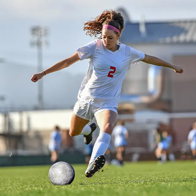 Female soccer player wearing a white jersey and black cleats kicking the ball on the field.