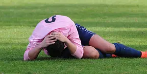 A soccer player lies on the ground, holding their head with both hands, likely due to an injury during a match. The player wears a pink jersey with the number 5 on the back, a blue jersey with shorts, and orange sneakers. The grass field is green and well-maintained, with some small leaves scattered on it. The sunlight illuminates the scene, casting shadows on the ground.