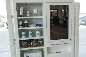 A white vending machine cabinet with a glass door is displayed, featuring a mirror on the door and a reflection of a person sitting on a chair at a table. Inside the cabinet, there are various items including bottles of eye drops and other products. Outside the cabinet, there is a blurry view of a parking lot with a car parked and a person standing, along with a car and a building in the distance.