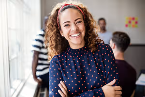 A woman in a blue polka dot top smiles at the camera in an office setting with people in the background
