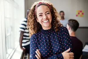 A woman in a blue polka dot top smiles at the camera in an office setting with people in the background