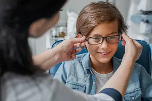 A young boy with glasses is having his ears checked by an adult woman, possibly in an optometrist's office.