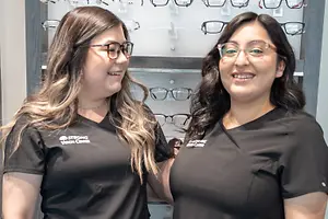 Two women wearing glasses and black t-shirts with the text 'Strong Vision Center' on them stand next to each other in front of a wall with glasses on display