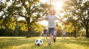 A young boy in a gray shirt and blue shorts kicks a soccer ball on a grassy field with other kids and trees in the background.