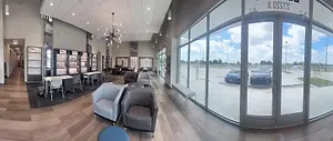 A panoramic view of a waiting room with chairs, tables, and glass walls, showcasing a modern interior design with natural lighting and a view of cars and trees outside.