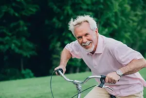 A man with a white beard and mustache rides a bike through a grassy field on a sunny day