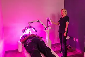 A man is lying on a white chair getting a light therapy treatment from a woman in a pink room