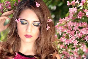A woman with long brown hair and colorful eye makeup poses in a garden with pink flowers.