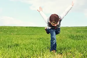 A boy with a baseball cap and sneakers jumps in the grass with his arms and legs spread out.