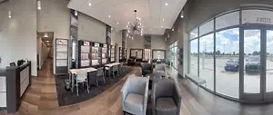 An image of the interior of a building with tables, chairs, and glass walls. There is a reception desk with a trash bin in front of it. There are also glass display cases with items on them. There are also some cars parked outside the building.