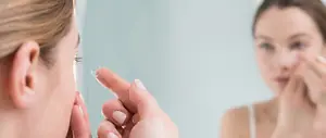 A woman is putting in a contact lens while looking in a mirror.