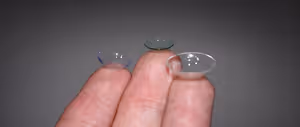 Two contact lenses are held between two fingers on a gray background.