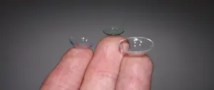 Two contact lenses are held between two fingers on a gray background.