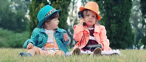 Two children sitting on a grassy field, one wearing a blue hat and the other a yellow hat, with the girl holding a toy microphone