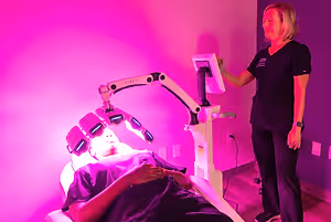 A woman is lying on a bed under a pink light, and a woman is standing beside her holding a monitor. The woman lying down is wearing a purple shirt, while the woman standing is wearing a black shirt with a logo on it. There is a pink wall with a power outlet and a plug in the background.